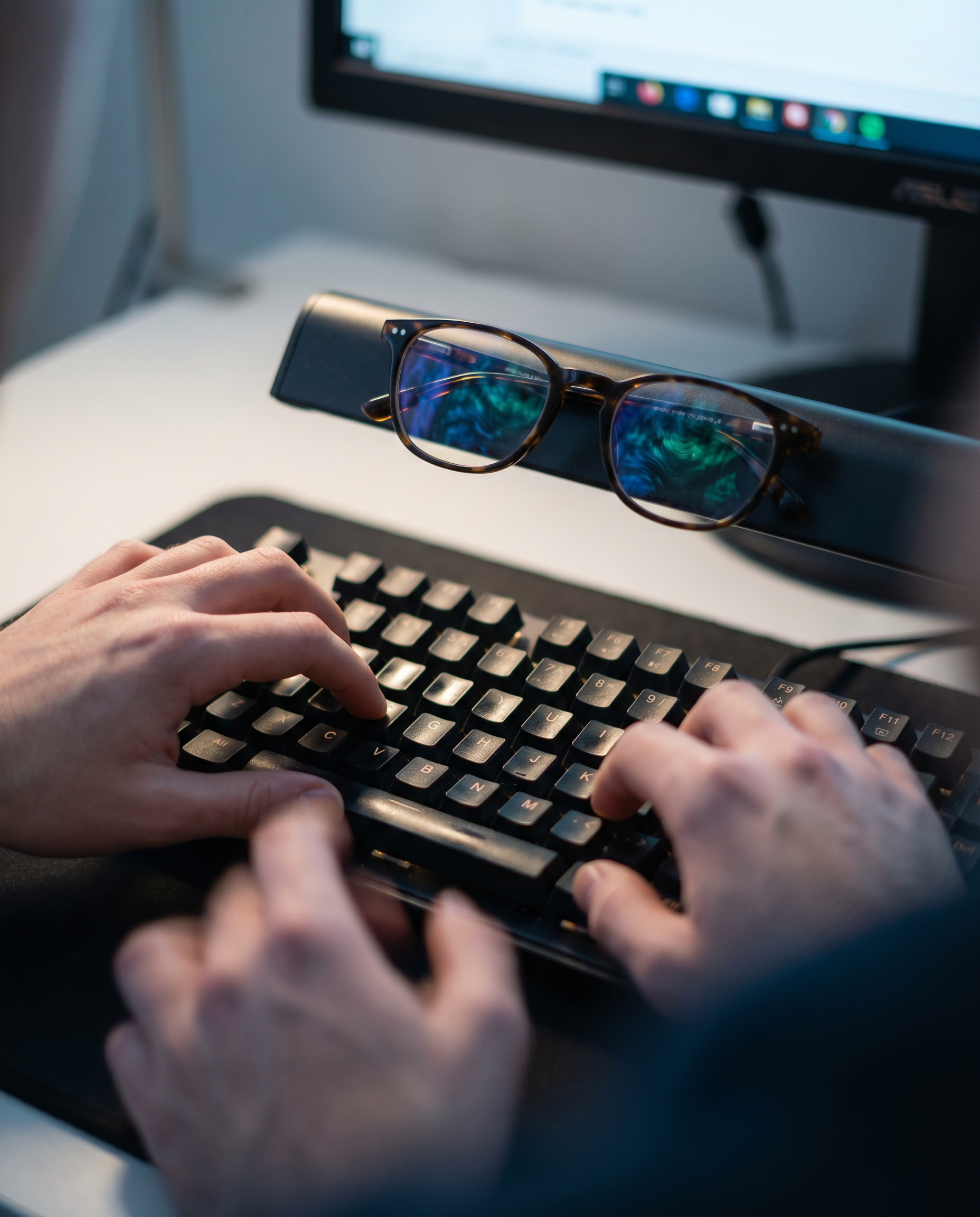 Photorealistic editorial close-up: human hands hovering over mechanical keyboard mid-typing, screen reflection visible in reading glasses showing abstract data visualization patterns in blue and green