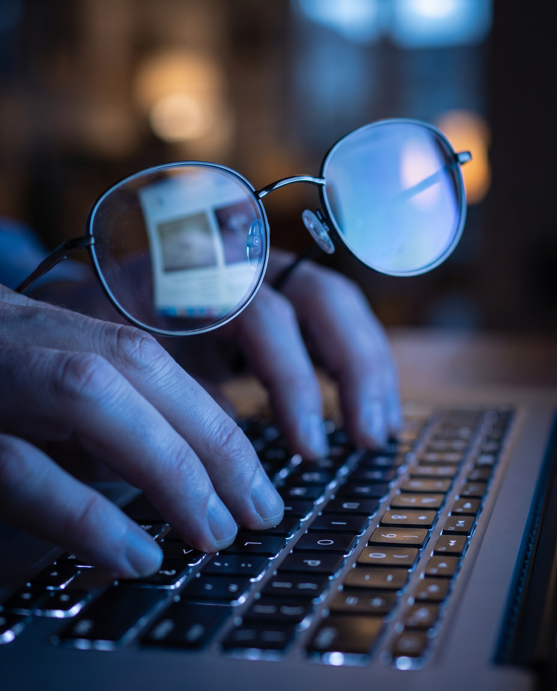 Photorealistic editorial photograph: extreme macro close-up of laptop keyboard with fingers typing, screen reflection visible in reading glasses showing split view of raw AI output text versus polishe