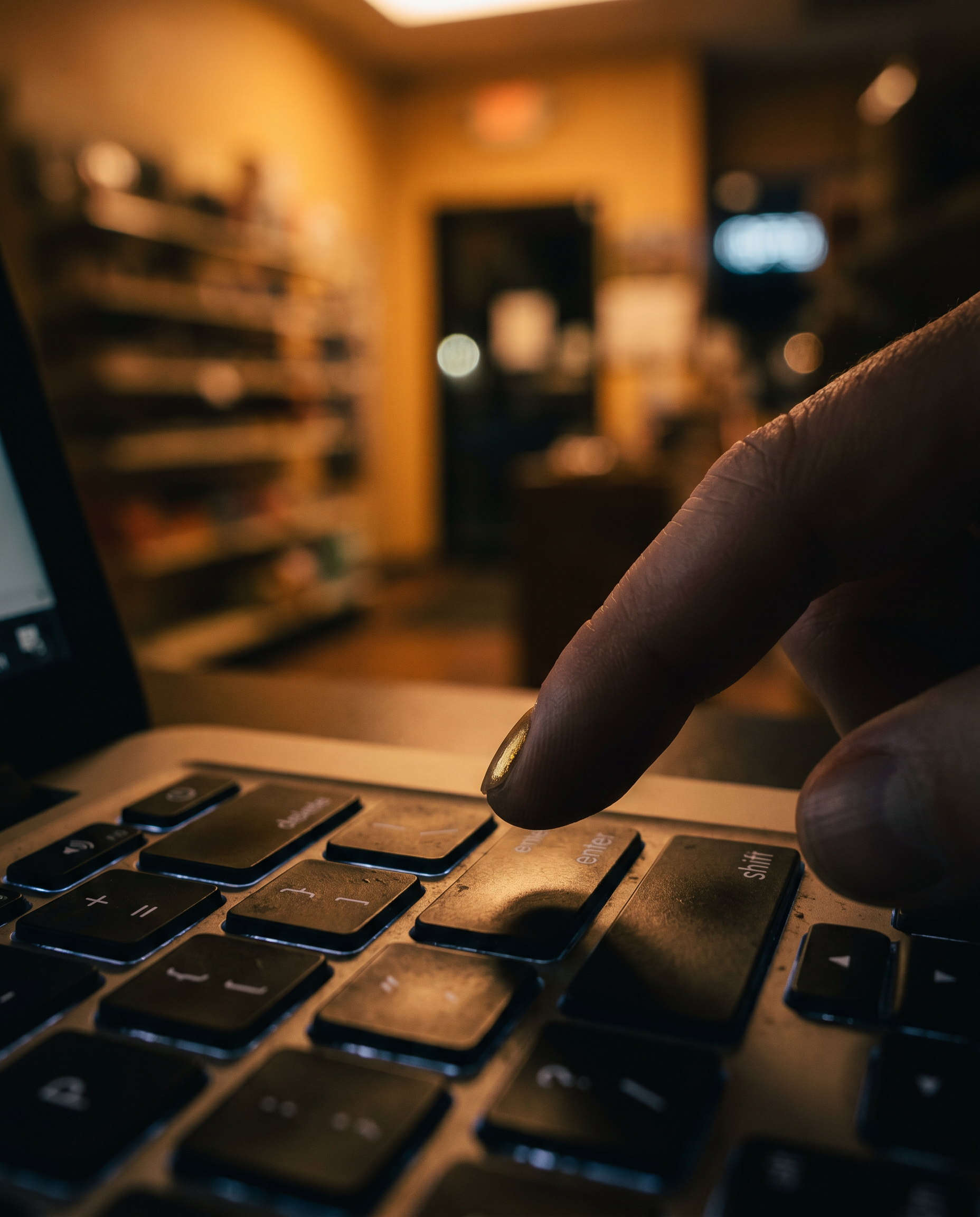 Photorealistic editorial photograph: dramatic close-up of a laptop keyboard with a single index finger hovering above the enter key, background an out-of-focus small retail shop with warm amber shelf
