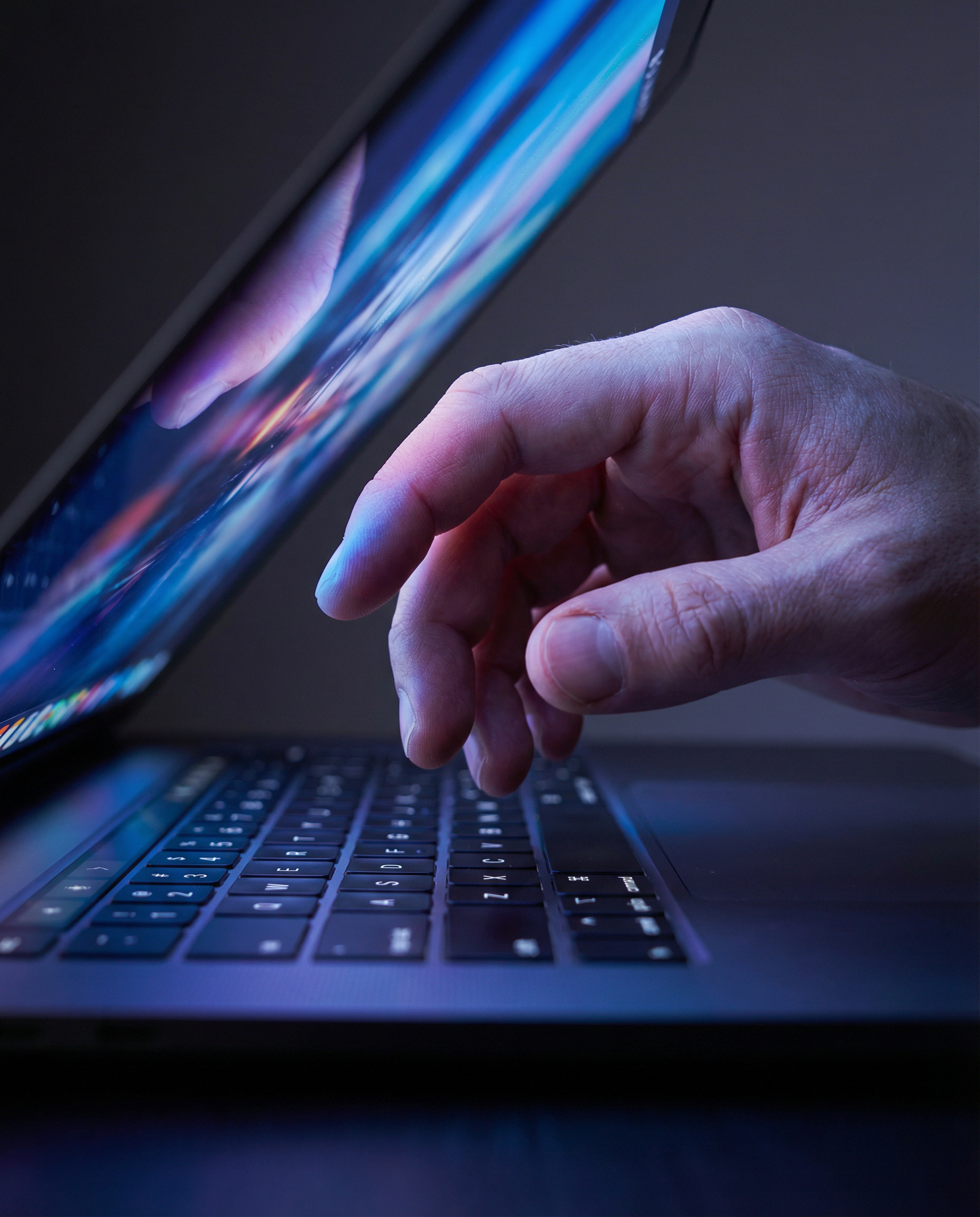 Photorealistic editorial close-up: human hand hovering hesitantly above laptop keyboard, screen reflecting colorful AI-generated text scrolling rapidly, fingertips caught mid-decision whether to edit